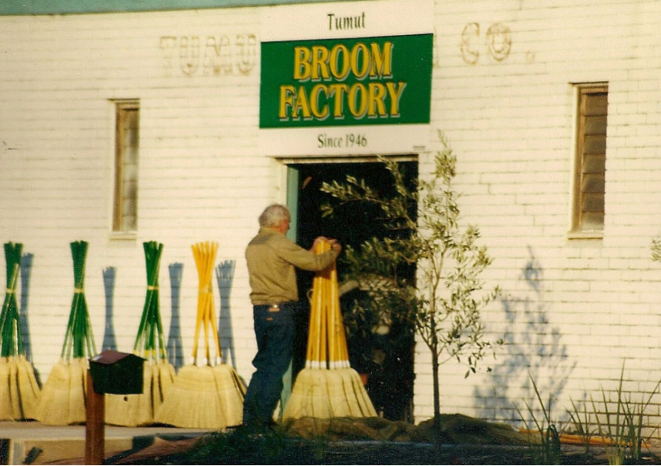 Vintage photo of a man with brooms in front of a building with 'Tumut Broom Factory' sign.