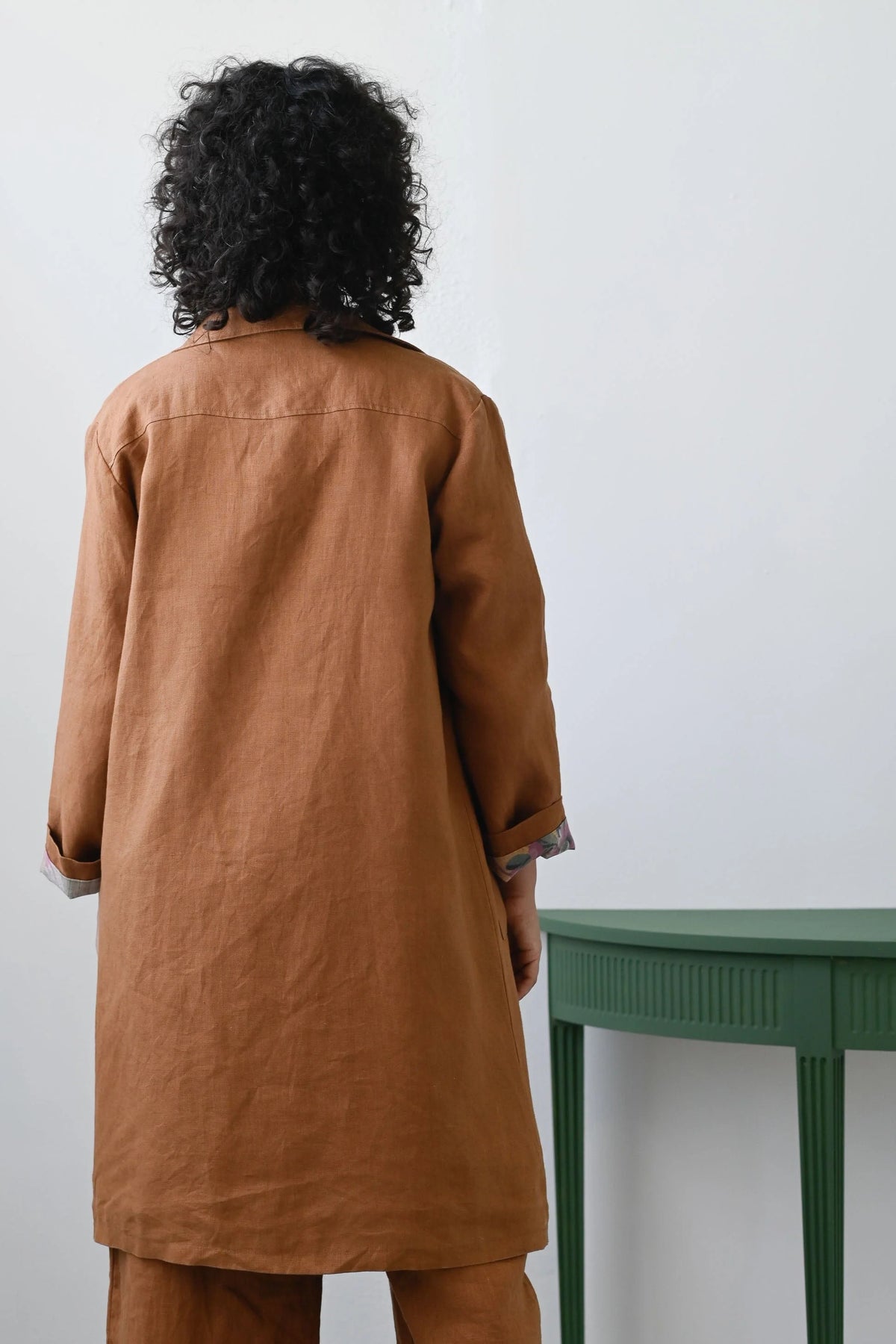 Person wearing a brown coat standing in front of a white wall with a green table.