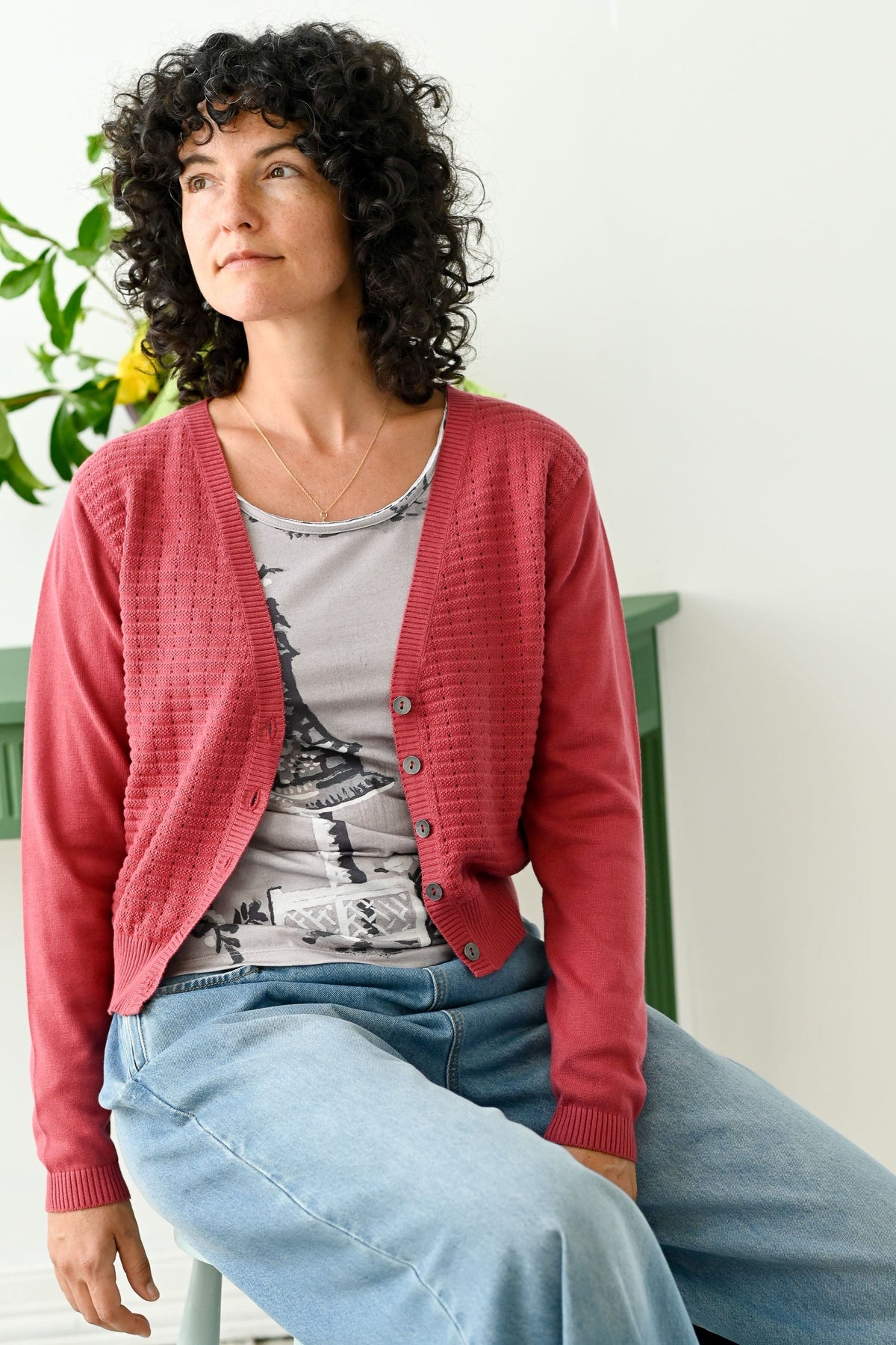 Woman wearing a pink cardigan over a patterned shirt and blue jeans, sitting on a green chair.