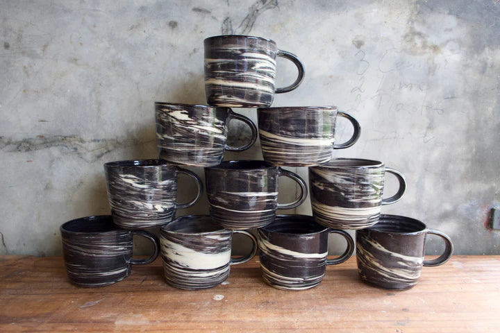 A stack of ceramic mugs with a black, white, and gray marbled pattern, arranged on a wooden surface against a textured concrete wall.