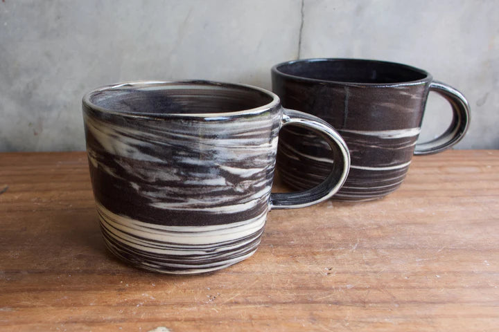 Two handmade ceramic mugs with a dark brown, black, and cream marbled swirl pattern, displayed on a rustic wooden table against a neutral gray wall.
