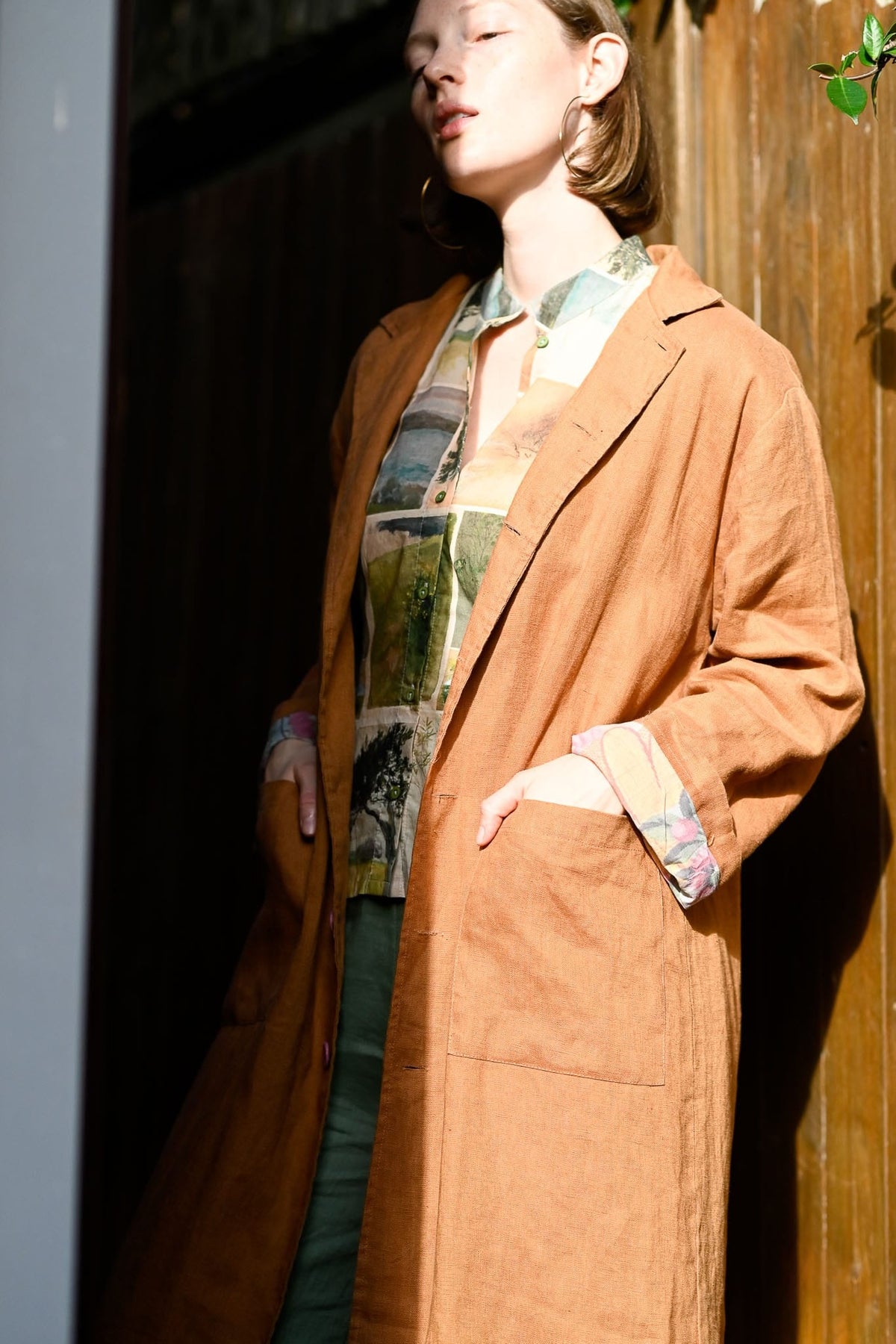 Person wearing a long brown coat with a plaid shirt underneath, standing against a wooden wall.