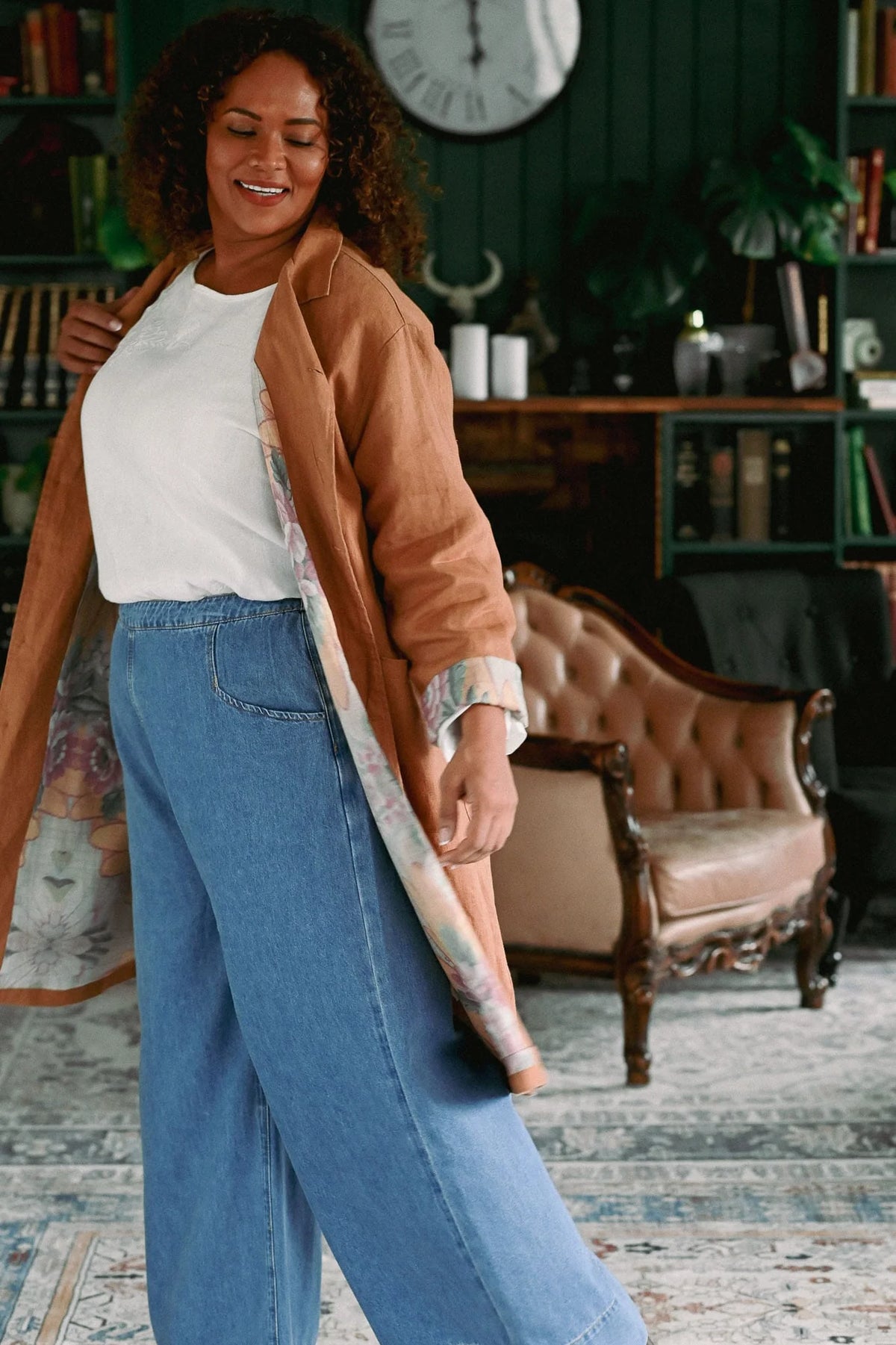 Woman in a living room wearing a brown jacket and blue jeans.