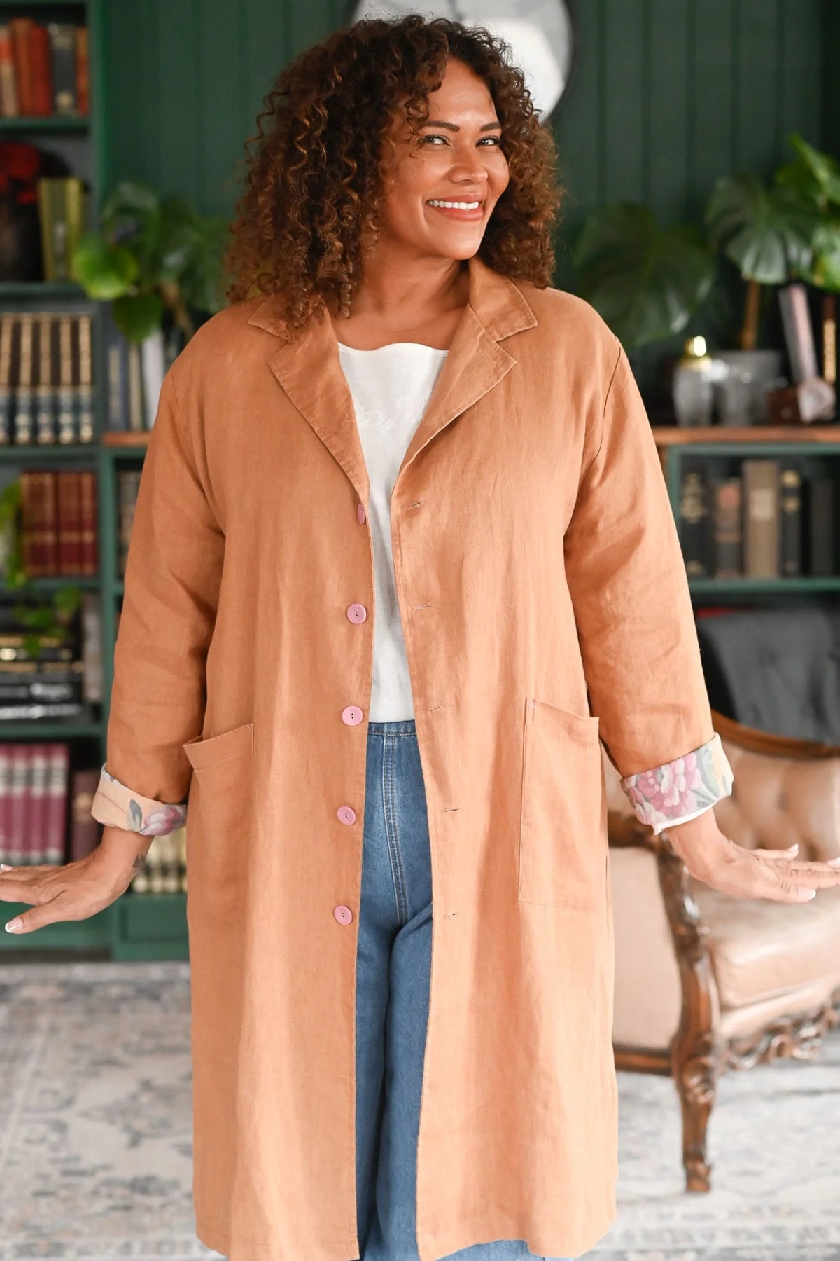 Woman wearing a long beige coat in a room with bookshelves and furniture.