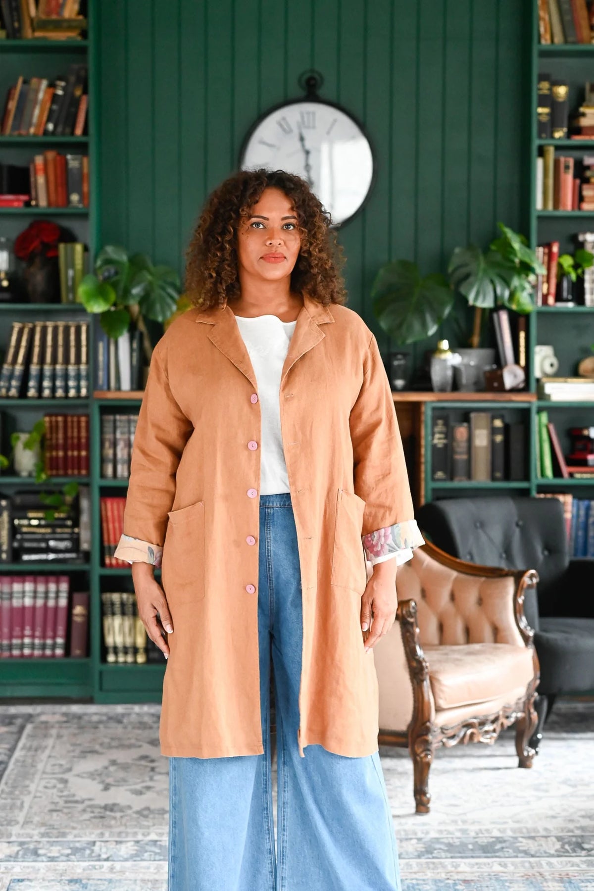 Woman in a beige coat standing in a room with bookshelves and a clock on the wall.
