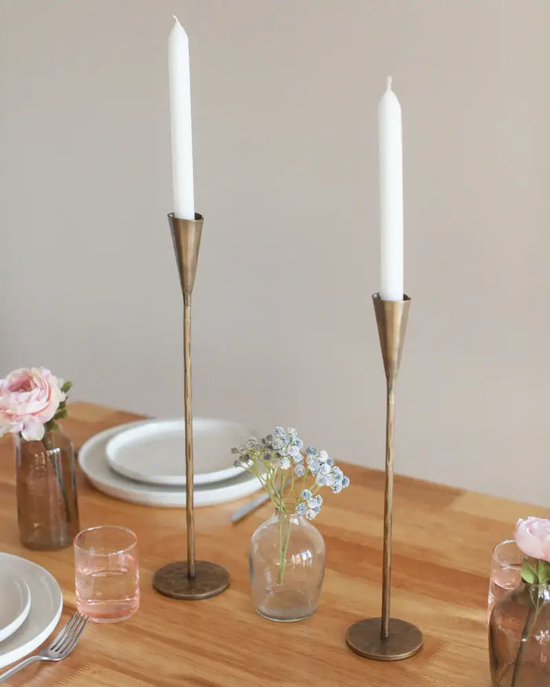 Two brass candle holders with white candles on a wooden table with plates and glasses.